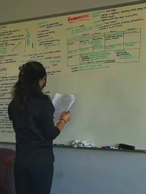 A woman stands in front of a whiteboard covered in handwritten notes and diagrams. She is holding a paper in her hands and is looking down at it. The whiteboard has the title "Biomolecules" and contains various notes and a table. The overall composition is a medium shot.