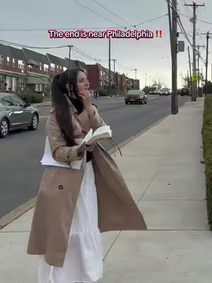 A woman stands on a sidewalk, holding a book and papers, with a street and buildings in the background. Text overlayed reads, "The end is near Philadelphia !!"