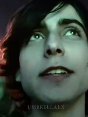 A close-up shot of a person looking up, with dark hair and fair skin. The lighting is dim. The background is dark, enhancing the focus on the person's face. The individual's expression is one of looking upwards, maybe focused on something above the camera.