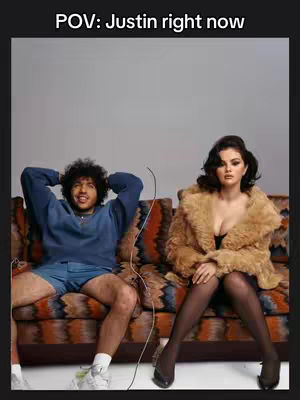 The image features a man sitting on a patterned couch, with another person, presumably Selena Gomez, seated next to him. Text at the top, "POV: Justin right now", sets a humorous context. The overall tone is playful and lighthearted, suggesting a relatable scenario.