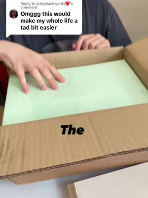A person interacting with an open cardboard box. Inside, there is a light green paper, and the person's hands are visible, reaching to touch it. At the bottom of the box, there is a tan/beige item, and the video is shot from a close up range.