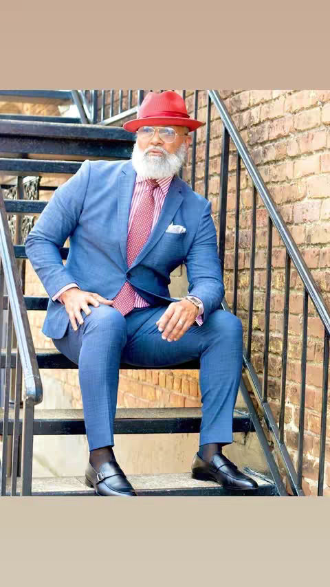 A man in a stylish suit is seated on outdoor metal stairs. He wears a blue suit, a red hat, and glasses. The background comprises a brick wall and the black metalwork of the stairs. The composition is formal and the lighting is bright.