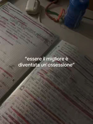 An overhead shot shows two open notebooks with handwritten notes, red underlining, and highlighting, resting on a dark surface. A pair of white earbuds, a colorful lanyard, and a blue bottle are visible in the blurred background. The text overlayed on the image reads "essere il migliore è diventata un'ossessione."