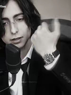 The image is a monochrome close-up of a young man. He is facing towards the left, holding a microphone up to his mouth. His right hand is raised, with a watch visible on his wrist. In the background, there is a glimpse of what could be a room setting. The overall composition is focused on the man's face and expression.