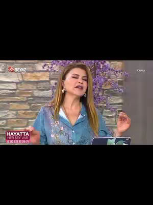 A woman with blonde hair and wearing a blue top with a floral design is the central focus of the image. She is positioned centrally with a subtle smile. A brick-like patterned wall serves as the backdrop. There are some purple flowers to the right. At the bottom left is a sign with text, and the "BEYAZ" logo is at the top left. The overall composition is balanced, conveying a studio setting.