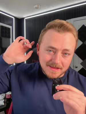 The cover image features a man with light skin, light brown hair, and a trimmed beard, positioned close to the camera. He wears a dark blue long-sleeve shirt. He has his hand close to his ear, suggesting he is listening or speaking. The background includes a black wall with vertical lines, and a portion of a white door frame, and a light fixture. A bottle of Coca-Cola is present on a table in the periphery.