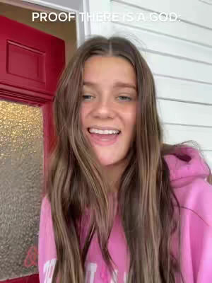 A young woman with long brown hair smiles widely at the camera, seemingly outdoors, with a red door and white siding in the background. The image has a bright, cheerful feel.