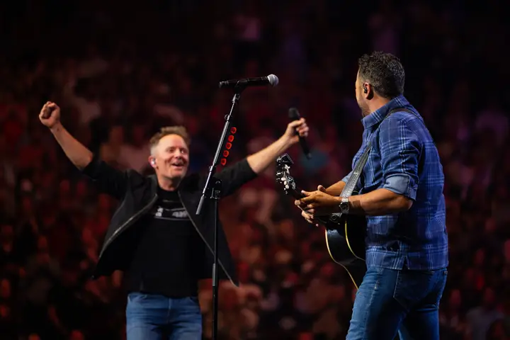 Two men on a stage with microphones, one with arms raised in excitement, the other playing a guitar. The background is blurred with many people.