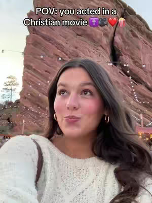 A young woman with long dark hair and wearing a white sweater faces the camera, looking upward. An outdoor background of a red rock formation and a cloudy sky is visible. Text overlay on the image reads "POV: you acted in a Christian movie" with appropriate emojis.