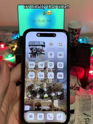 A person's hand holding a smartphone displaying a Christmas-themed homescreen, which is held up in front of a laptop and a cup, all placed on a surface with Christmas lights. The phone's screen features custom app icons and widgets, enhancing a festive vibe. The background includes Christmas decorations, the laptop screen displaying festive-themed background and other festive elements.