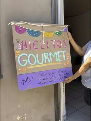A handmade sign advertising 'Hielitos Gourmet' is held by a person's hands. The sign is made of a brown cardboard background with colorful decorations and text. The text is in Spanish, advertising gourmet ice pops with various flavors and a price.