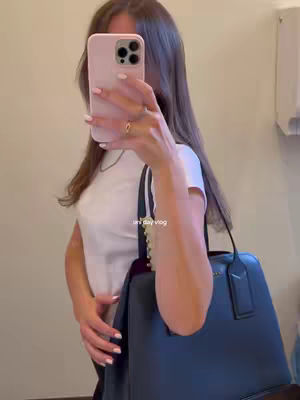 A woman is taking a mirror selfie with a pink phone, holding a dark blue handbag. She is wearing a white t-shirt and a silver necklace. The background is a light-colored wall. The image is well-lit, with a modern and clean composition.