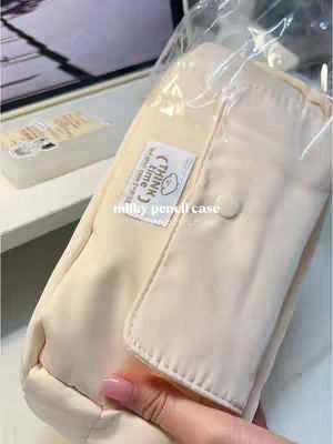 The cover image shows a cream-colored pencil case in a clear plastic bag. A hand with pink nail polish is holding the pencil case. The pencil case is rectangular with a flap closure and a small label that reads 'think time' and 'milly pencil case'. The background is blurry, with hints of a desk and possibly a laptop.