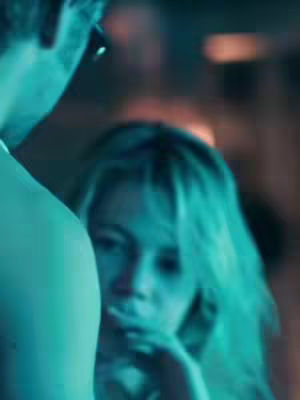 The image shows a close-up of a woman's face partially obscured by another figure. The woman has blonde hair and appears to be looking at something off-camera with her mouth closed. The lighting has a blue-green hue.