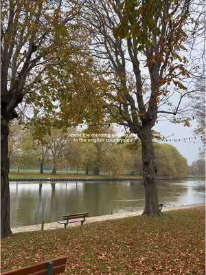 A serene landscape shot featuring a tranquil lake with trees lining the banks on either side. The trees display autumnal foliage, and a bench sits near the water's edge. The sky is overcast, giving muted colors to the scene.