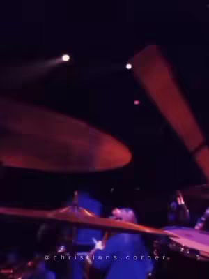 An artistic, dimly lit shot appears to be taken at a concert. The composition features the top of a drum set lit by dynamic lighting: a large, curved cymbal overhead, with a drummer obscured in the foreground. Red and white spotlights play across the dark background, giving the impression of a live music environment.
