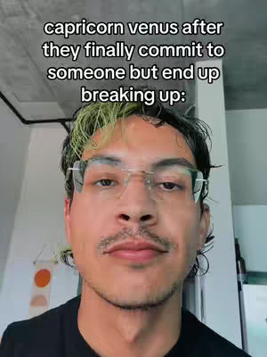 A selfie video shows a man in a room, looking directly at the camera. He has dark hair with a dyed green streak and wears glasses. Text overlayed reads: capricorn venus after they finally commit to someone but end up breaking up:
