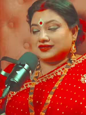 The close-up shot features a woman with dark hair, a red dot bindi, and gold earrings; she is wearing a red sari with gold accents and a gold necklace. She is holding a microphone for a potential broadcast or recording.