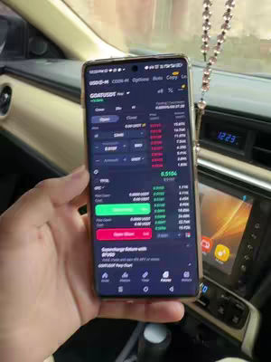 A hand holding a smartphone displaying the Binance app interface. The app shows trading charts and order information. The background subtly reveals the interior of a car.