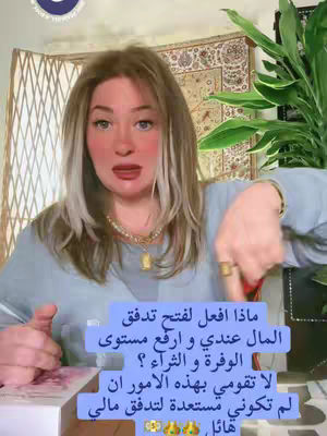 A woman with long blonde hair and fair skin is centered in the frame, looking directly at the viewer with a slight smile. She wears a light blue top, jewelry with a pendant, and a gold ring. A block of blue text, in Arabic, is superimposed over the lower portion of the image, adjacent to her hands.