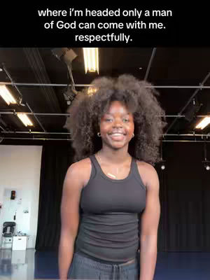 A young woman with dark curly hair smiles at the camera in a studio setting. She wears a black tank top and has a gold necklace. The background includes studio lights, black curtains, and a dark floor. Text is overlaid at the image.