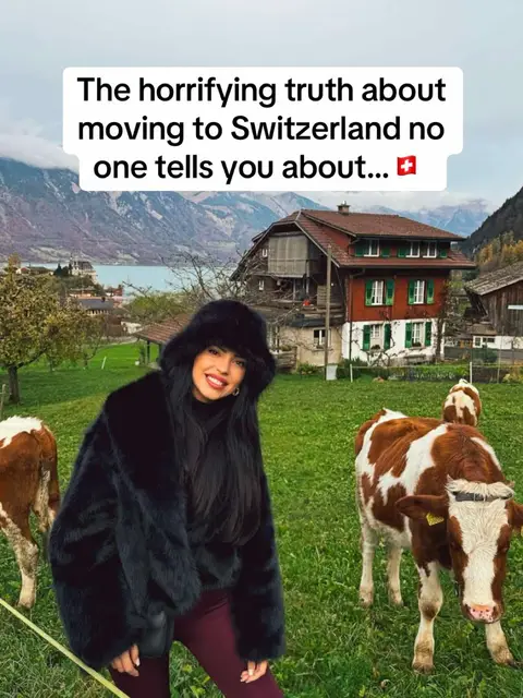 A woman in a black fur coat and hat smiles in front of a scenic Swiss landscape with cows and a house.