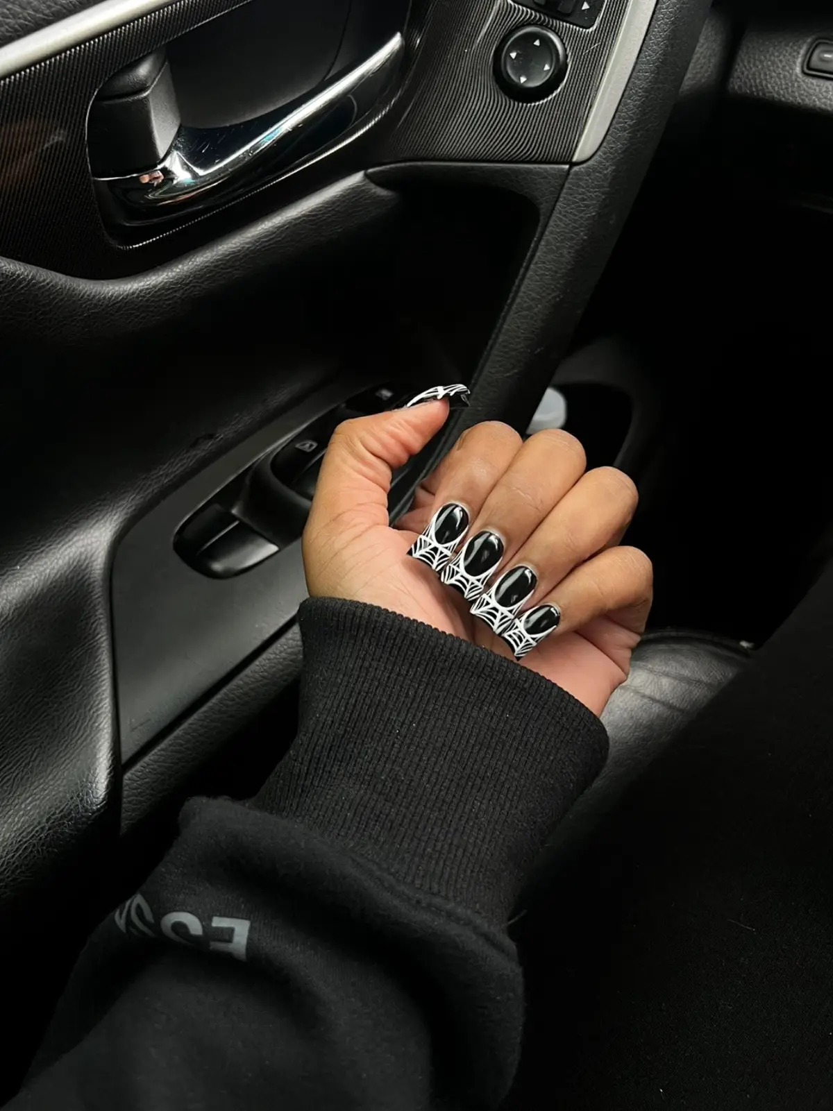 The image shows a close-up of a hand with long, black, and white nail art. The hand is posed inside a car, with the car's interior visible in the background, including a door handle and window controls.