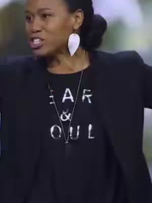 A medium shot features a dark-skinned woman speaking with emphatic expression. She has her hair up in a bun and is wearing a black blazer over a black t-shirt. Her large, white, leaf-shaped earrings are prominent, as is a silver necklace hanging over the shirt. The shirt has the words "FEAR & SOUL" printed on it. The background is blurred.