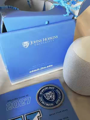 Image showing a light blue box and brochure from Johns Hopkins University. Displayed is the university’s logo, the words “Johns Hopkins University” are visible. A commemorative coin and a white speaker are also parts of the picture.