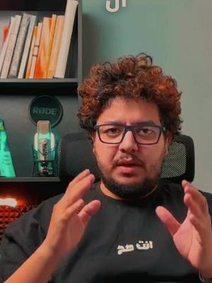 A man with curly brown hair and glasses is seated in front of a shelf of books and a microphone, with his hands raised in a gesture of explanation. The background is a mix of a green wall and a shelf of books. He's wearing a black t-shirt with Arabic text. The lighting appears to be natural, possibly from an overhead source.