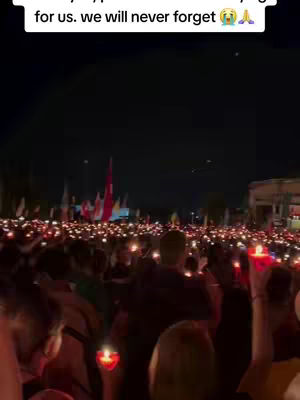 An outdoor shot at night, showing a crowd of people holding candles. The scene is predominantly dark with numerous small, flickering lights creating a warm glow against the darkness. In the background, there are blurred structures, flags, and more lights.