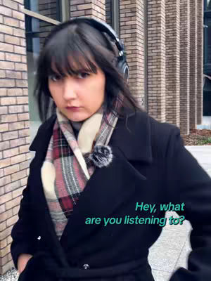 The cover image shows a woman with dark hair wearing headphones. She is outdoors, standing in front of a building with brickwork. She wears a dark coat and scarf with a plaid pattern, and the overlay text suggests the video's content.