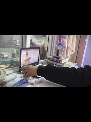 A desk is set up with a laptop showing a video call. A  person is holding a pen over papers. A lamp and stack of books are visible.