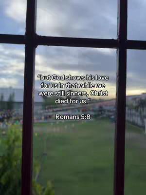 The image shows a view through a window frame. The sky is overcast with soft, muted colors. The text overlay features a quote from Romans 5:8, set against a backdrop of a green field and buildings in the distance.
