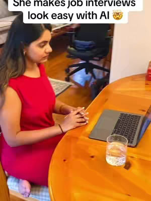 A woman in a red dress sits at a round wooden table with a laptop and a glass of water, a bright window is in the background, emphasizing the setting with natural lighting.
