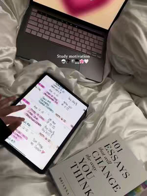 The image shows a laptop and a tablet placed on a bed with white sheets. The laptop is open, displaying a blurred pink image. The tablet shows a study schedule. A book titled "101 Essays that will change the way YOU THINK" is placed to the right of the tablet. The overall tone is neat and focused.