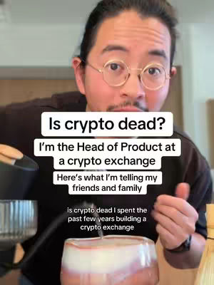 A man with glasses and a mustache is standing in a well-lit space, holding what appears to be a cocktail glass filled with a white and reddish liquid. Overlay text in white boxes asks if crypto is dead and introduces the speaker as the Head of Product at a crypto exchange.