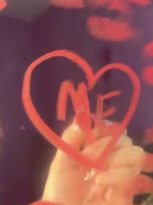 A hand holding a pane of glass is seen with the word "ME" scrawled inside a heart shape. The heart is drawn in bold red, while the glass appears to be reflecting a purplish background highlighted with blurred red circles.