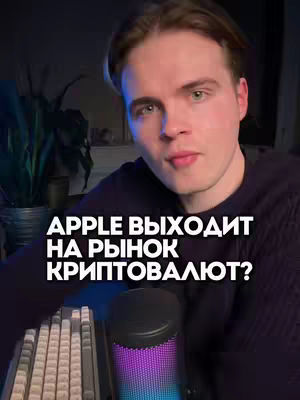 A young man with light skin is the central focus, set against a dimly lit background. Overlayed on the image is bold, white text asking if Apple is entering the cryptocurrency market. The man has short, brown hair, and he gazes directly at the viewer. The background hints at a home setup with a green plant and a glowing microphone situated near a keyboard.