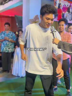 A man in a white t-shirt is holding a microphone, looking slightly downward and to his right. Behind him, other people are visible in a brightly lit room, possibly at an event. The text overlay says "EH IKAW?"