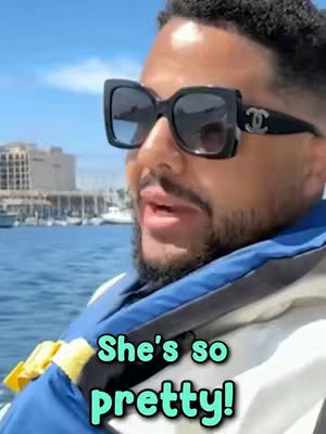 A man wearing large sunglasses speaks, with a teal and white outlined text box conveying the words 'She's so pretty!' in a bubble-like font, with a background showing bright blue sky and water.