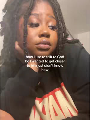 A selfie-style image features a young Black woman with long, dark, locs. The background is blurred, suggesting an indoor setting. The woman is wearing a dark, long-sleeved top with a portion of a bright red and yellow design at the bottom, possibly a logo. Her facial expression is pensive, with her chin resting on her hand and her eyes looking downward. Text is overlaid on the image.