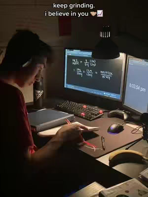 A young man is sitting at a desk, studying late at night. Two computer monitors are in front of him, one displaying mathematical equations. A notebook and pen are on the desk, alongside a water bottle and other items. The lighting is dim, creating a focused, studious atmosphere.