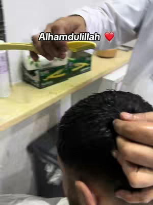 The image shows a close-up of a person's head with dark hair as a barber shaves the hair off the top of the head using a yellow razor. The text overlay "Alhamdulillah" appears above the scene, with a red heart symbol next to it, indicating a sense of gratitude or completion. The background includes a shelf with boxes on it.
