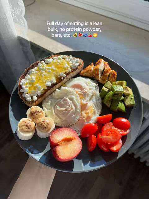 A top-down shot of a plate of food featuring a variety of healthy items, including toast with a yellow spread, fried eggs, sliced chicken, avocado, tomatoes, and a cut plum. The plate is dark with patterns, set against a bright background, possibly near a window.