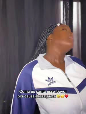 A woman with dark skin is seen from the chest up, looking upwards with her eyes closed, as if in song. She is wearing an Adidas athletic jacket in white, navy, and light gray, with her hair in braids. Text overlays at the bottom of the frame.