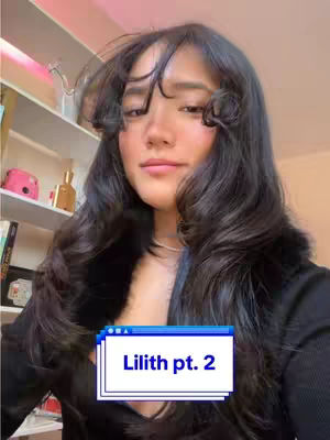 A woman with long, dark wavy hair looks directly at the camera. She wears a black top. A text overlay at the bottom reads 'Lilith pt. 2'. The background features a shelf with items.