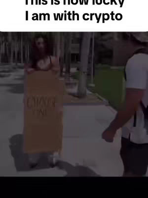A woman walking on a sidewalk holding a cardboard sign, with text overlaying. Another person is partially visible in the background.