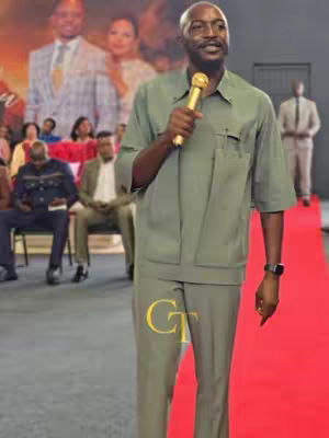 A man in a green suit stands center with a microphone, on a red carpet. The image has a backdrop of people and a blurred image of a couple. The color is a set of muted tones.