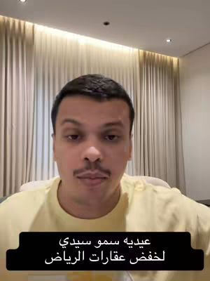 A man is centered in the frame, facing the camera directly. He has short, dark hair and a mustache. He wears a light yellow t-shirt, and his expression is neutral. Behind him, a softly lit curtain creates a warm backdrop, and a black banner at the bottom contains white text.